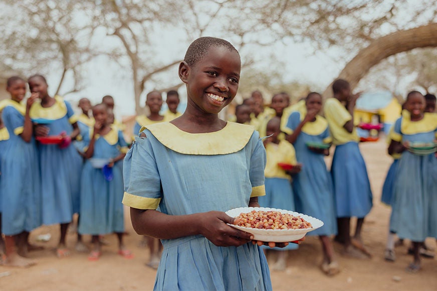 A girl in a school uniform holds a bowl of nutritious food. A group of classmates smile behind her.