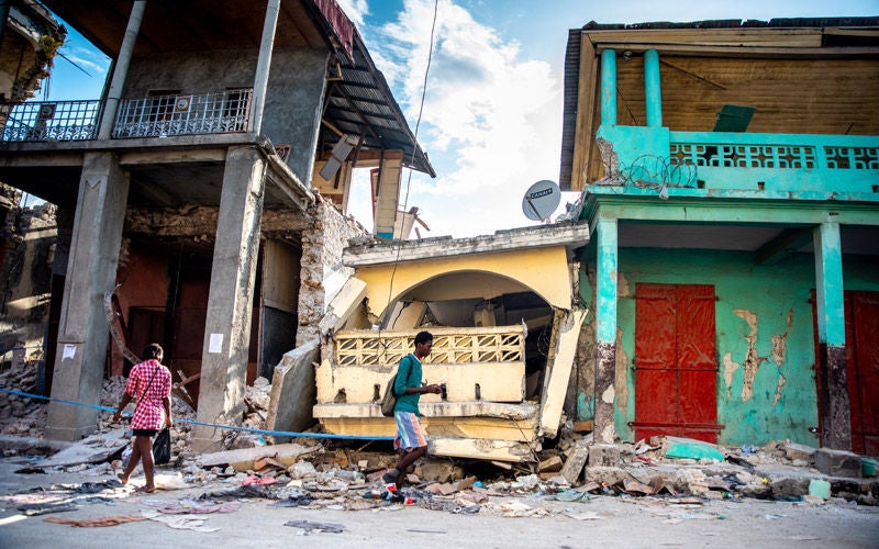 Two Haitian people walk in opposite directions down a street with brightly coloured ruined buildings behind them.