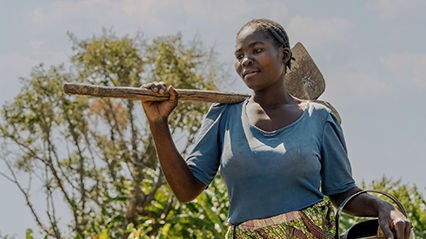 A woman in a blue shirt carrying a hoe and watering can outdoors, surrounded by greenery.