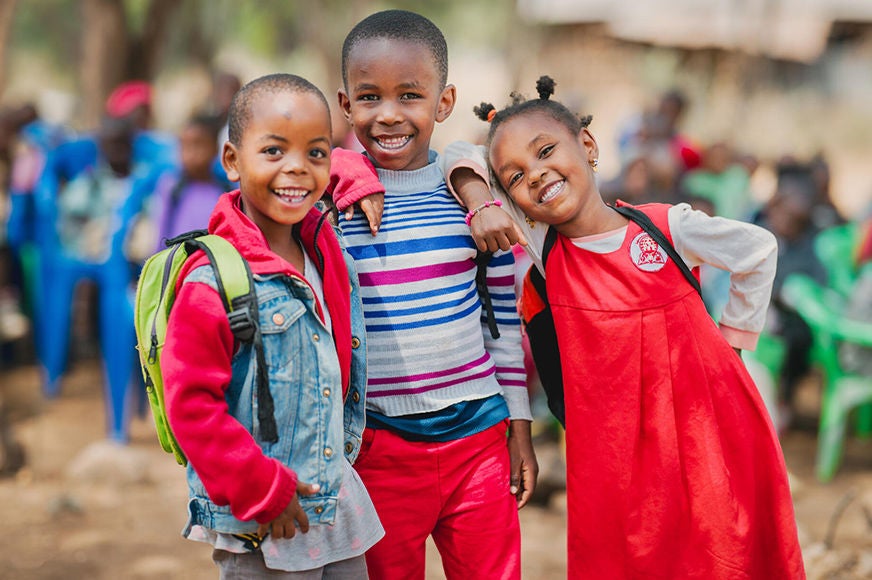 Three children with backpacks stand smiling outside their school. (Tanzania, 2023)
