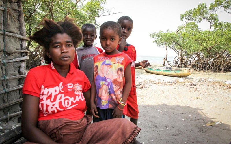 A woman sits outside a house made of mud and sticks. Three children stand near her.