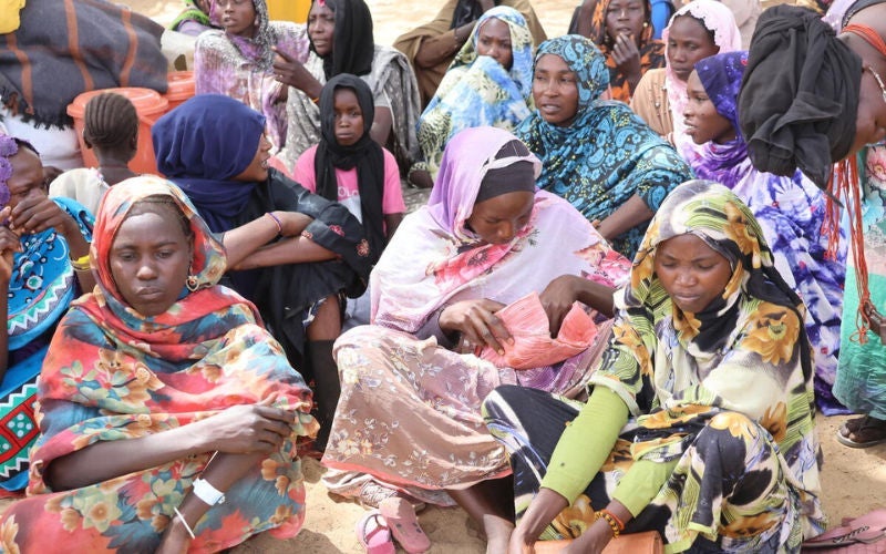 A group of people sit closely together outdoors, wearing colorful patterned clothing and head coverings.