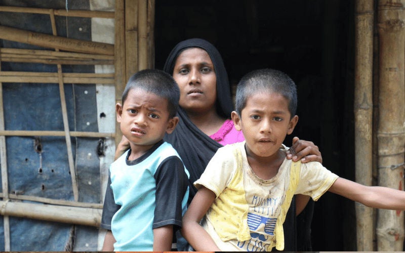 A woman and two school-age boys sit together outside a rough shelter.
