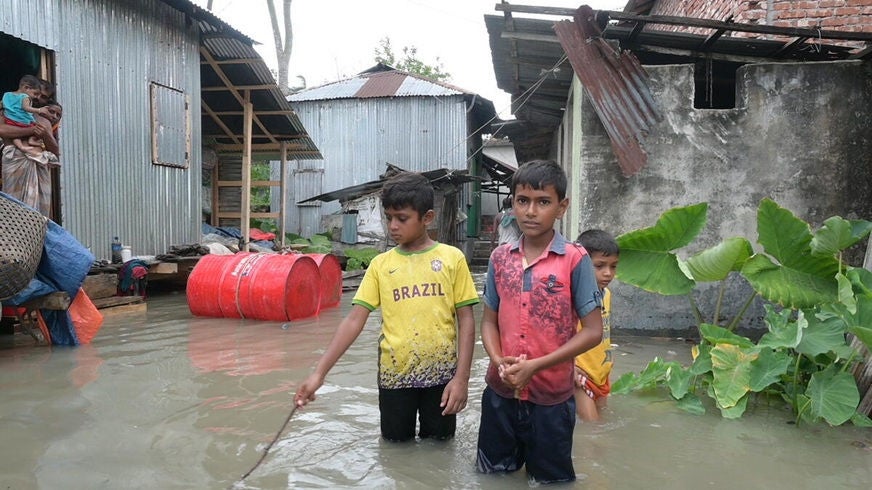 Children stand in knee deep floodwater between metal and concrete homes in a rural area. Debris, barrels, and large green leaves are visible around the waterlogged surroundings. (Bangladesh, 2024)