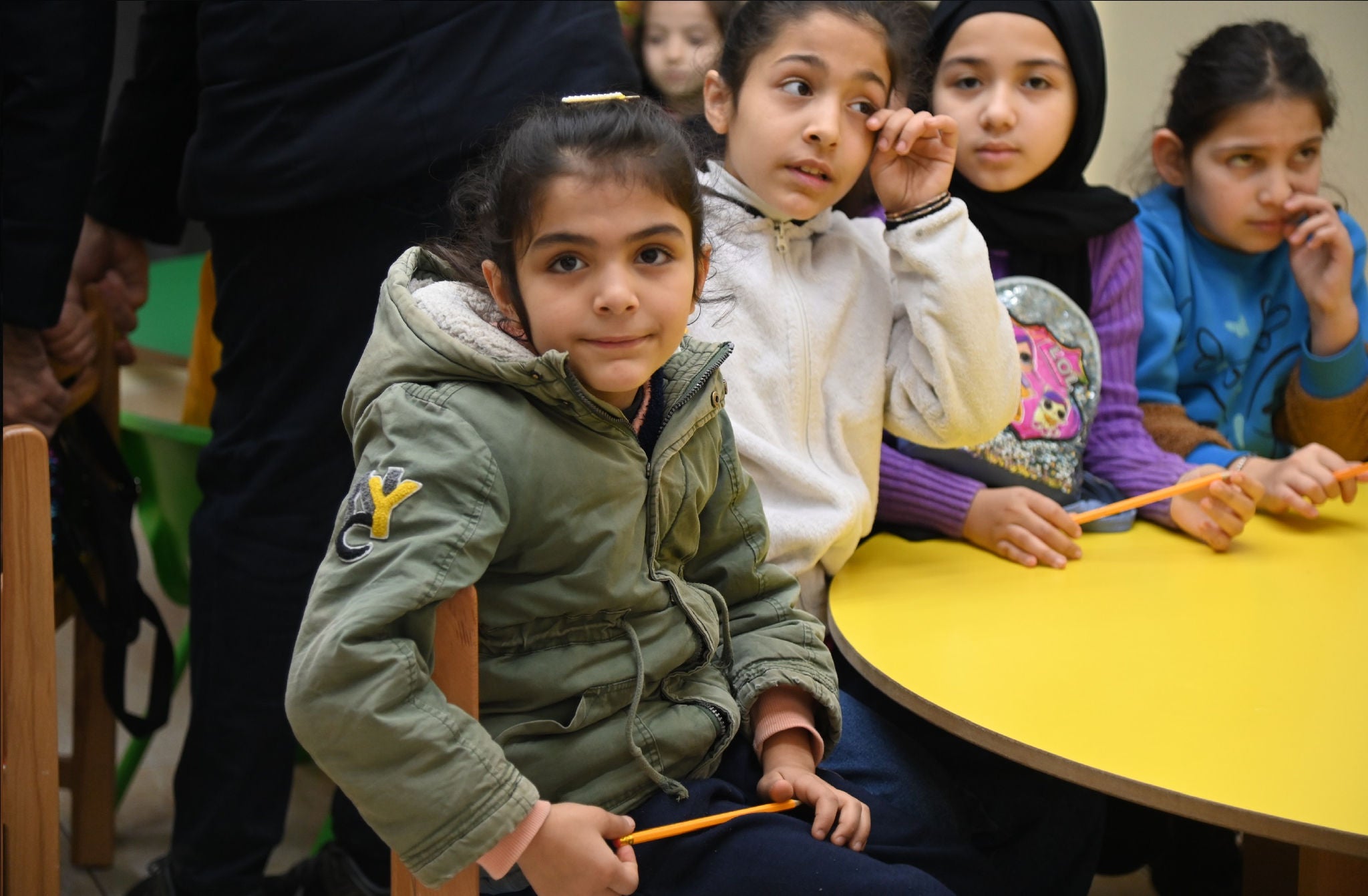 A group of children sit around a table, each child holding a pencil.