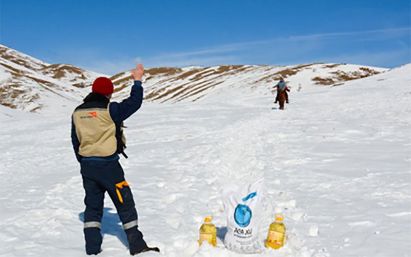 A World Vision worker in snow pants and a wool hat waves to a man riding an animal out of the mountains.