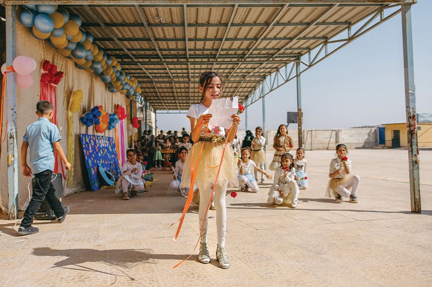 A young girl wearing a white shirt and yellow skirt is reading a letter after participating in a community event. (Syria, 2024)