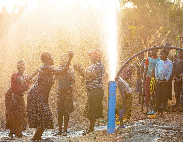 A group of people dancing and celebrating as water sprays into the air from a well.