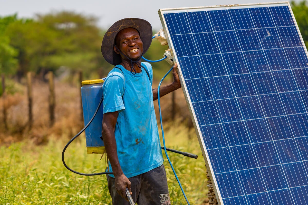 A person wearing a wide-brim hat and blue shirt stands in a grassy field holding a solar panel, with a backpack sprayer and hose visible. (Malawi, 2025)