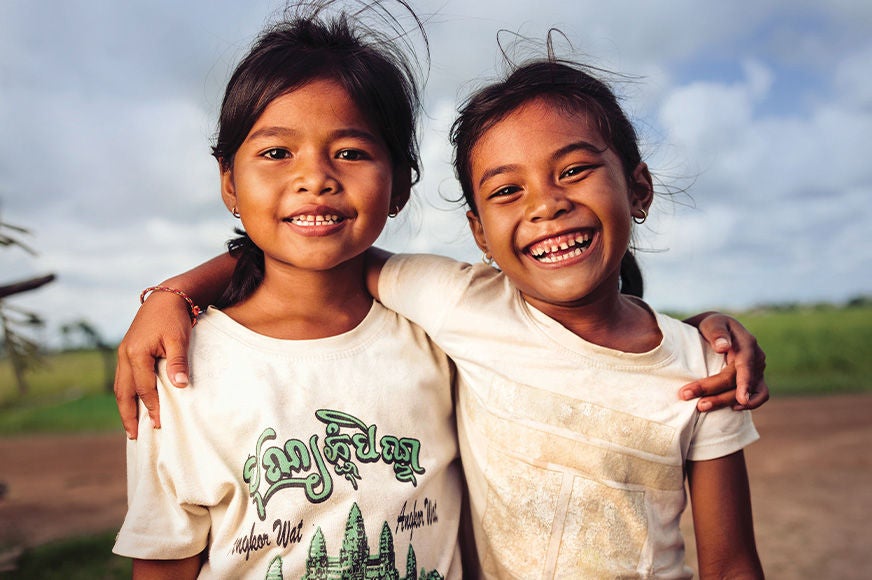 Two young girls wearing white t-shirts smile broadly as they stand side by side in a grassy field, their arms on each other’s shoulders. (Cambodia, 2025)