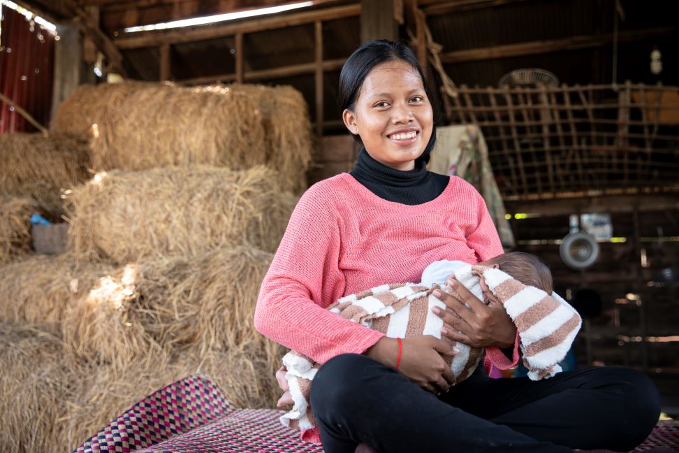 In Pouk District, Cambodia, a mother sits on a mat in a barn, nursing her infant.
