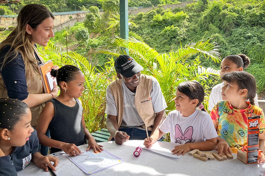 World Vision staff leading outdoor educational activity with children, featuring drawing materials and games on a table in a lush, green setting. 