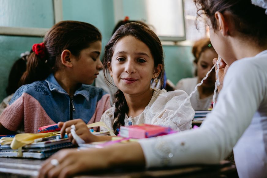 In Syria, a young girl in a busy classroom sits at a desk loaded with school supplies.
