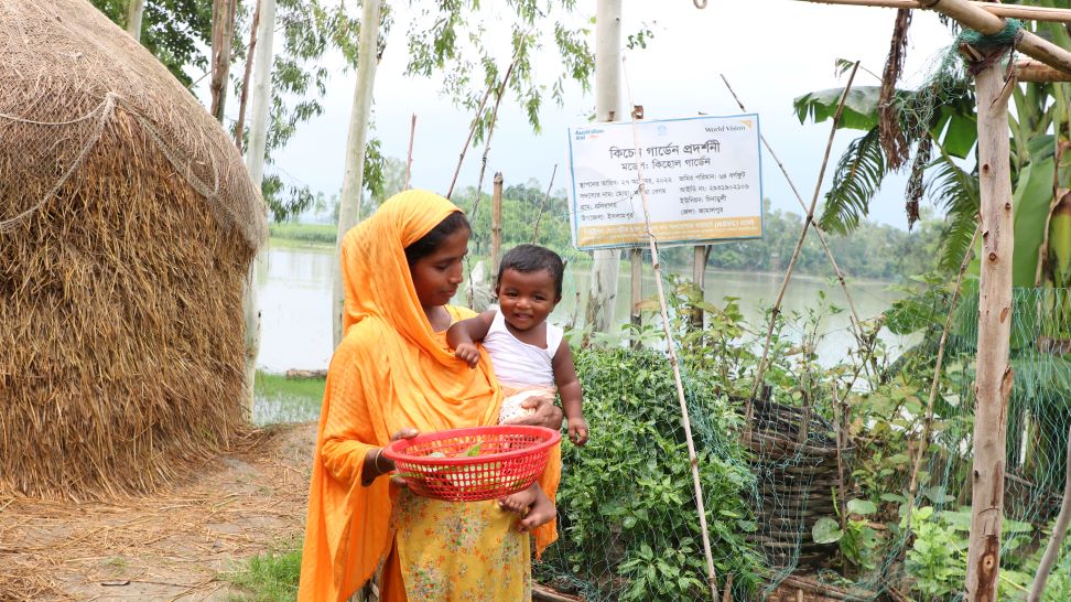 In Islampur, Bangladesh, a woman balances a baby on her hip while collecting garden produce.