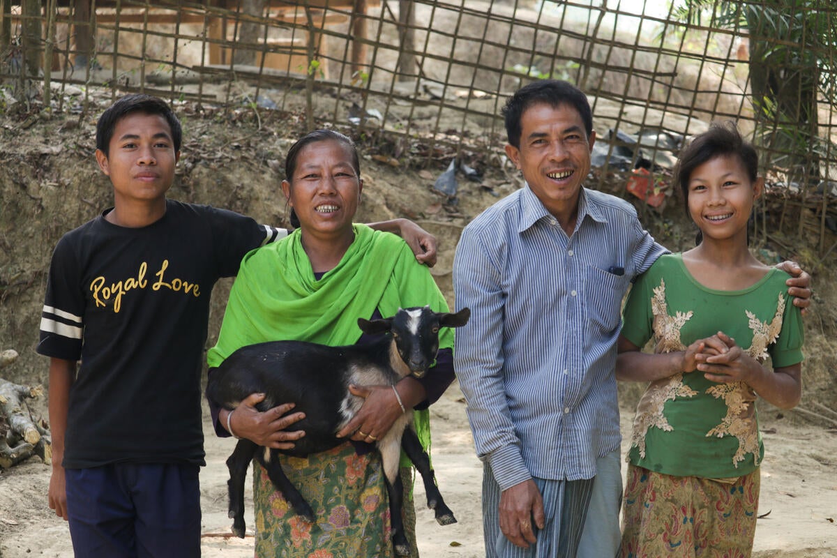 A family of four in Cox’s Bazar, Bangladesh stand smiling together. The mother holds a young goat.
