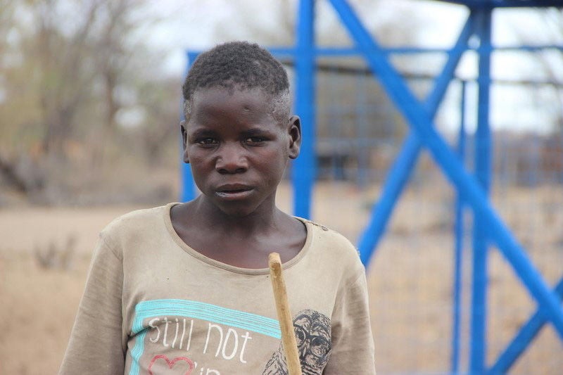 A teenage boy stands in a dry-looking environment, with a wooden tool after one arm.