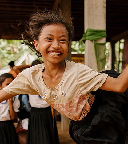A girl jumping mid motion is wearing a pink floral shirt, smiling widely.