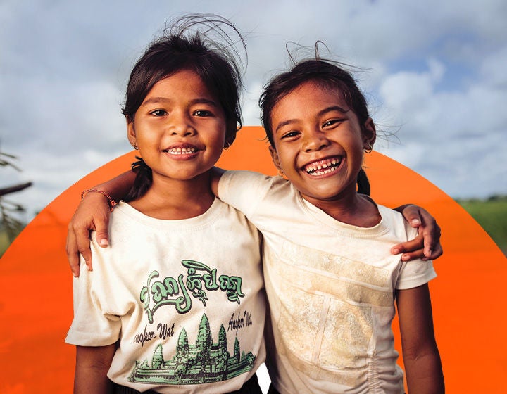 Two young girls wearing white t-shirts smile broadly as they stand side by side in a grassy field, their arms on each other’s shoulders. (Cambodia, 2025)