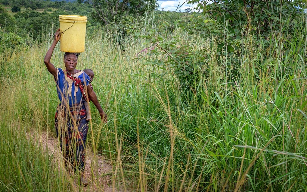 A woman carries her baby and a bucket of water on her head.