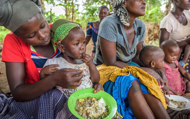 A group of mothers feed their young children from bowls, sitting on the ground.