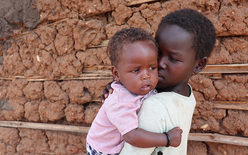 A young girl holds a baby girl in her arms as they stand in front of a mudbrick wall.