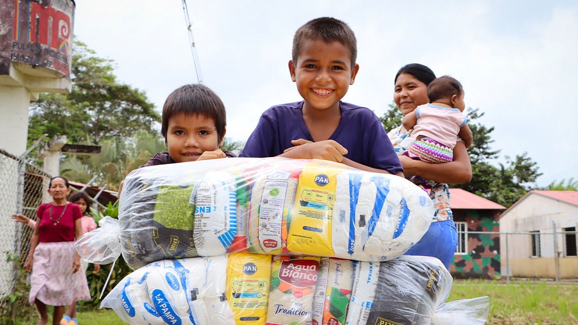 Two boys standing outdoors, smiles, while leaning over a large bundle of emergency food supplies. (Venezuela, 2025)