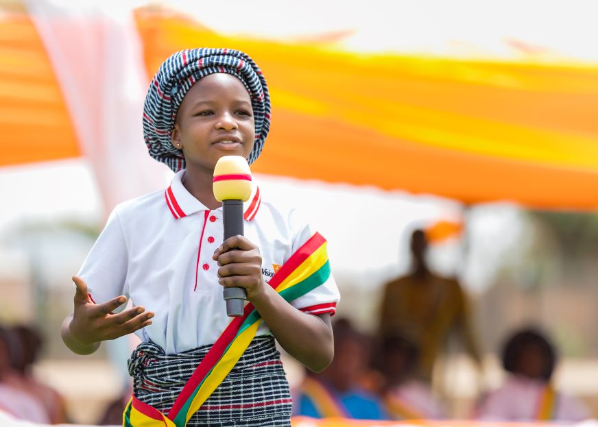 A child wears a World Vision polo shirt and sash, and speaks into a microphone.