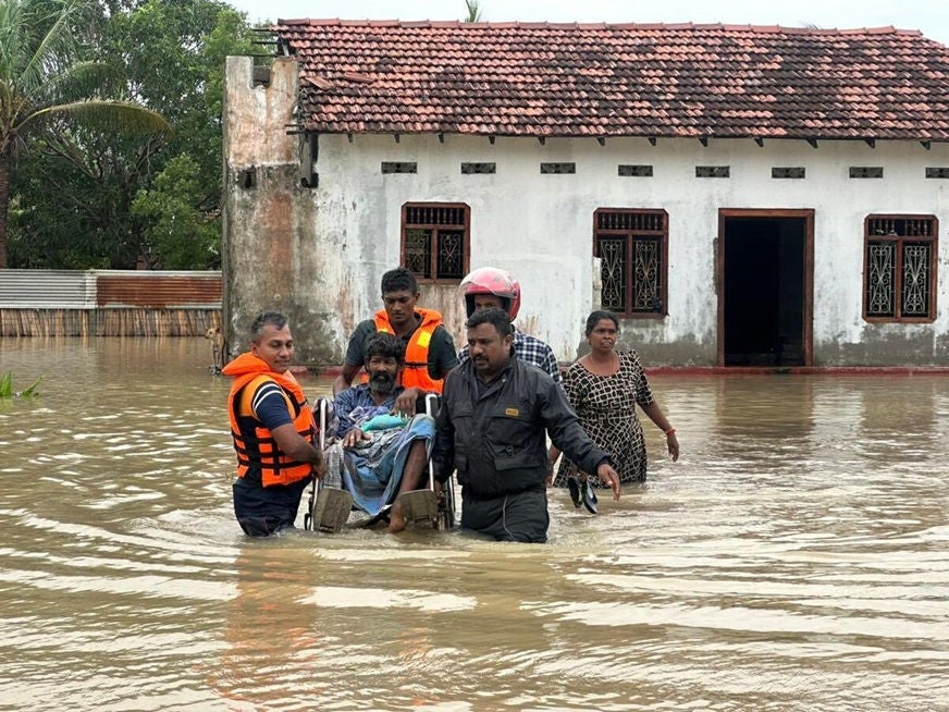 Four men and a woman wade through thigh-high water as they rescue a disabled man in a wheelchair. (Sri Lanka, 2025