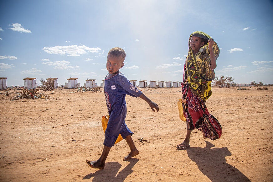 A young boy and girl carry water jerry cans across a vast, dry landscape under a bright, clear sky.