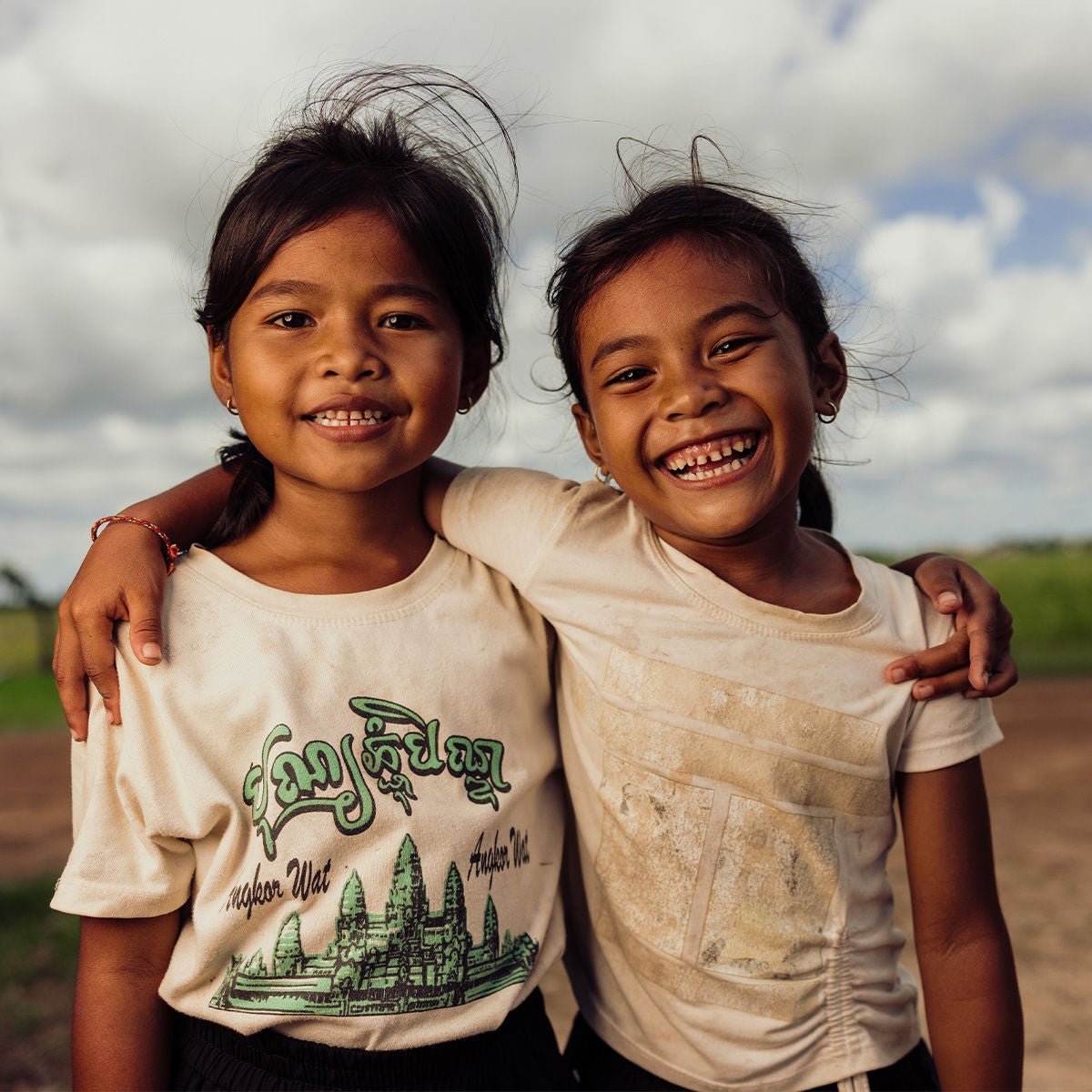 Two young girls stand with their arms across each other's shoulders, smiling.