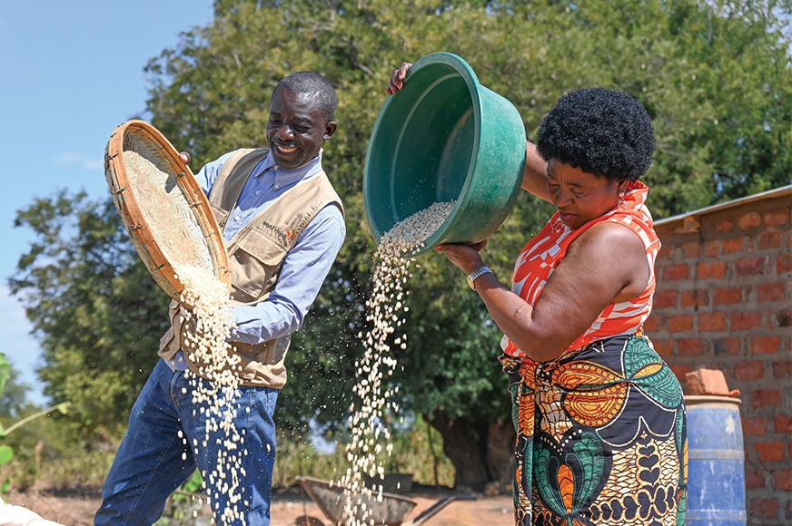 A man in a World Vision vest and a woman in a colorful dress pour grain from their vessels. (Zambia, 2024)
