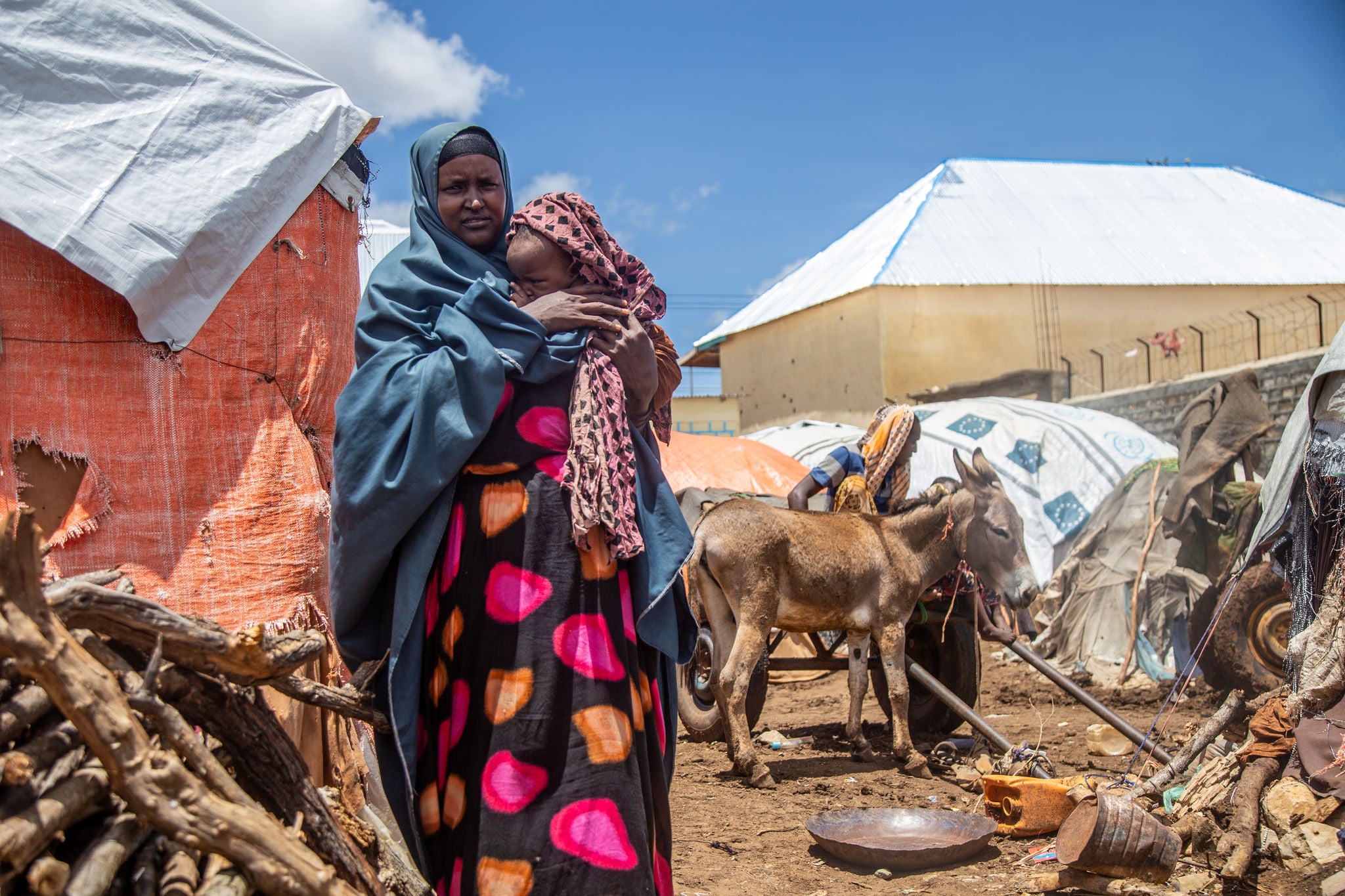 A woman in bright clothes and a head scarf holds a baby in front of her shelter. 