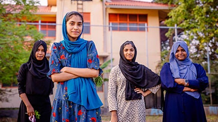 Four teenage girls are standing outdoors in front of a building with a red roof, surrounded by greenery.
