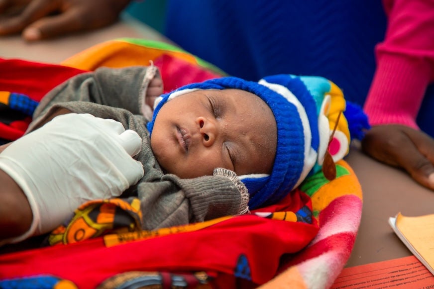 An infant sleeps in a blue hat while the gloved hand of a nurse conducts a medical exam. 