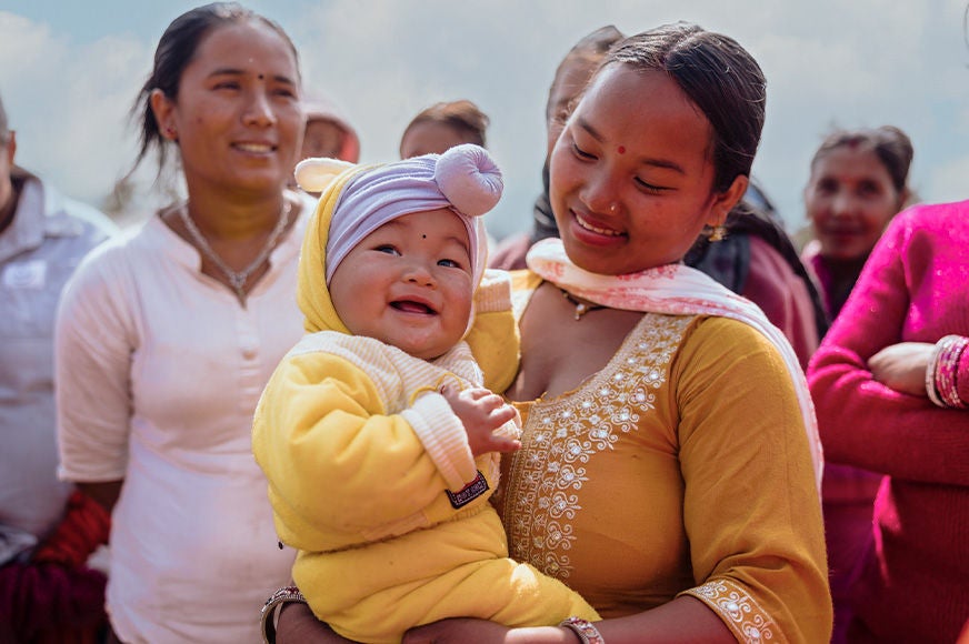 Mother holding her baby in a yellow outfit while standing among a group of women during a community gathering.