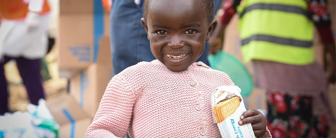 A young girl smiles, holding a high energy biscuit. (DRC, 2024)