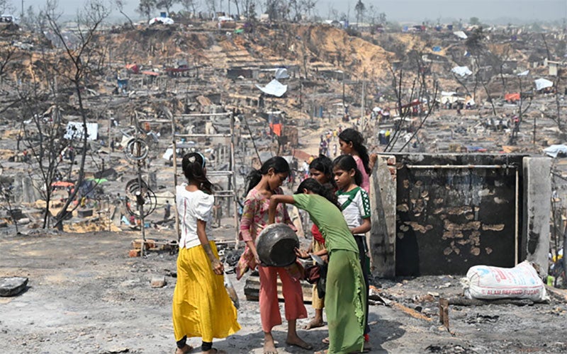A group of girls in Cox’s Bazar refugee camp hold and look into a bowl or cooking pot. Around them lie the charred remains of the camp.