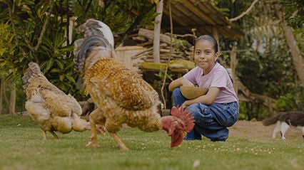 Young girl kneels behind her chicken with a bowl of feed.