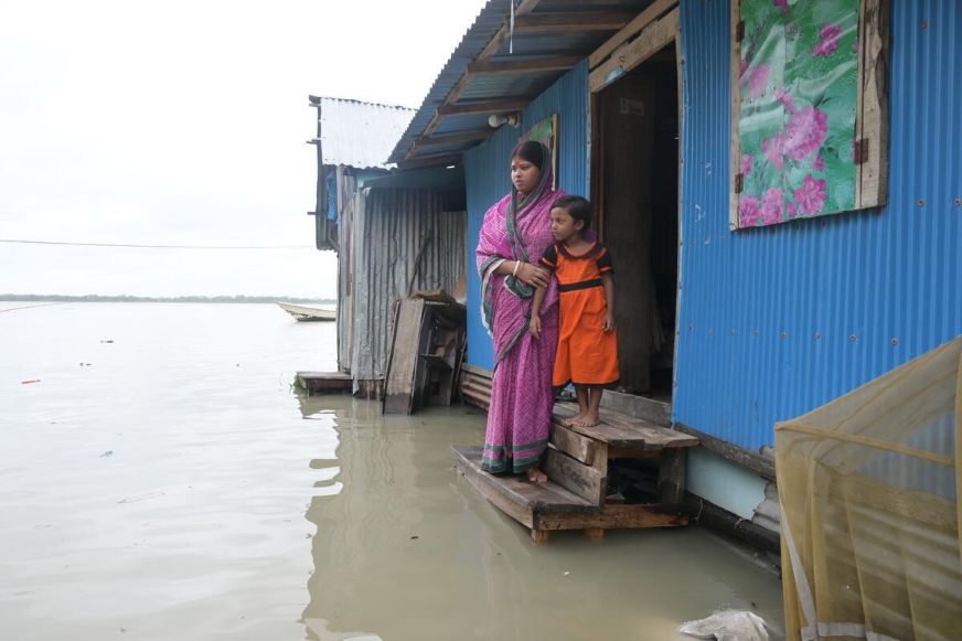 An adult in a pink patterned garment holds the arm of a child wearing an orange outfit. They stand on wooden steps outside a blue corrugated-metal house, staring worriedly at floodwater that surrounds them. (Bangladesh, 2024)