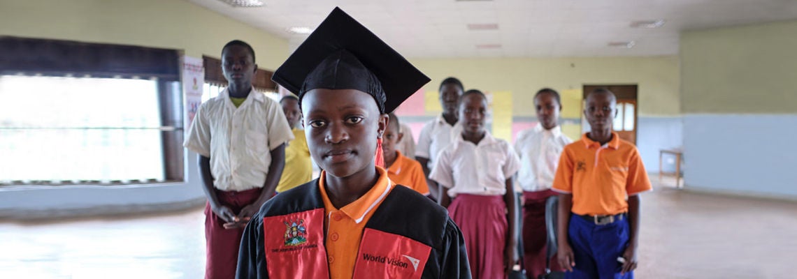 World Vision student in graduation gown with classmates in school uniforms inside a classroom setting.
