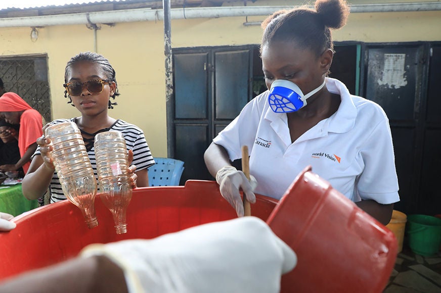 An adolescent girl wearing sunglasses pours two plastic bottles while another adolescent girl stirs.