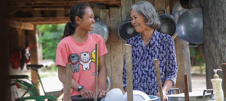 Two people washing dishes outdoors in a rural setting, with one wearing a pink shirt featuring cartoon characters and the other in a blue patterned shirt. Various kitchen utensils and pots are visible around them.
