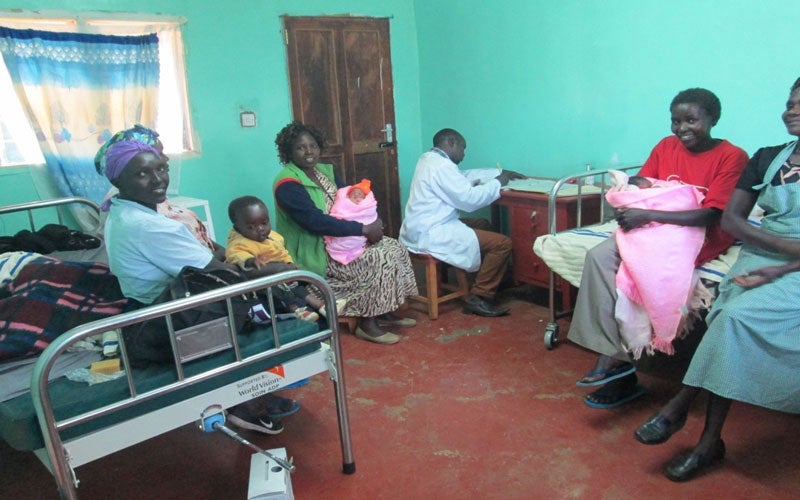 Holding their babies, a group of four women smile as they sit in a medical office while a health-care worker works at his desk.