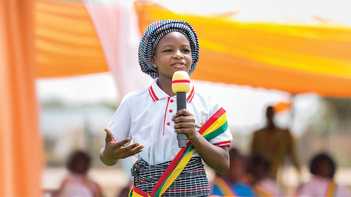 Child holding a microphone and speaking at an outdoor cultural event under bright yellow and orange canopies.