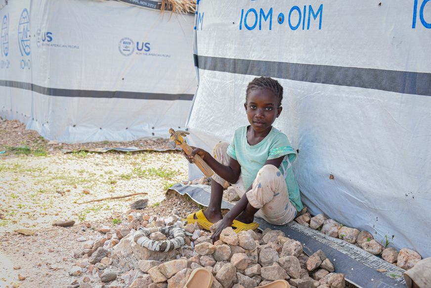 A young girl in Democratic Republic of Congo sits outside a temporary home, smashing medium-sized rocks into gravel. 