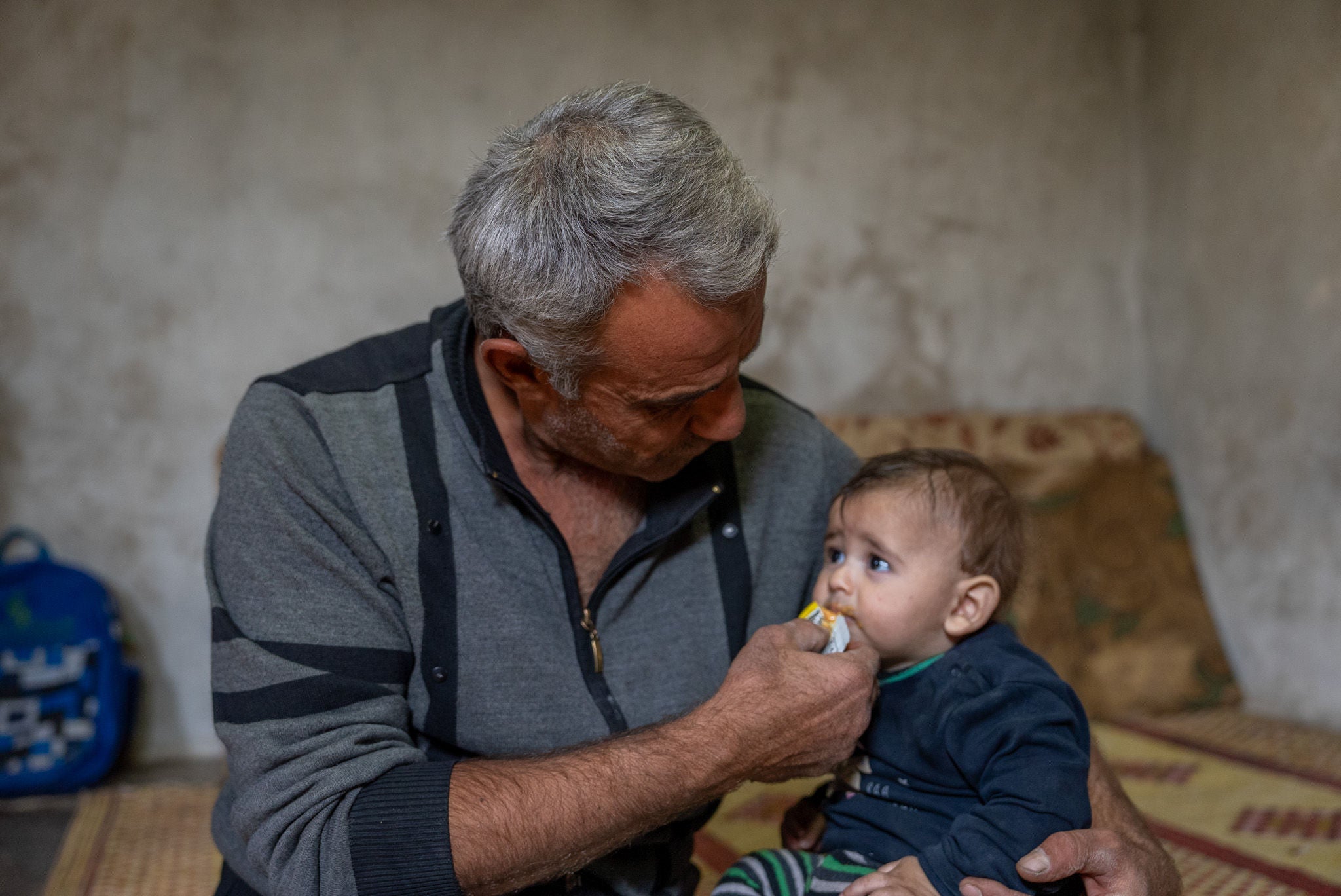 A father holds his infant on his lap, feeding a pack of therapeutic food. (Syria, 2024) 