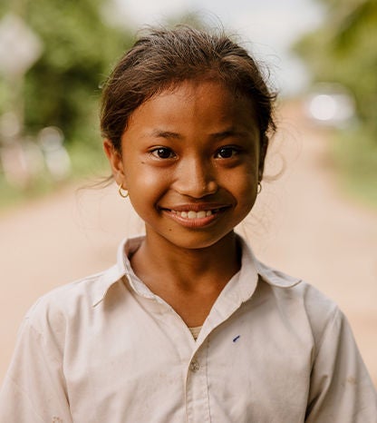 A girl wearing a white uniform stands on a dirt road smiling. 