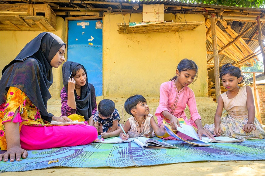 Children of various ages are doing their schoolwork on a blue mat as an adult woman watches over them.