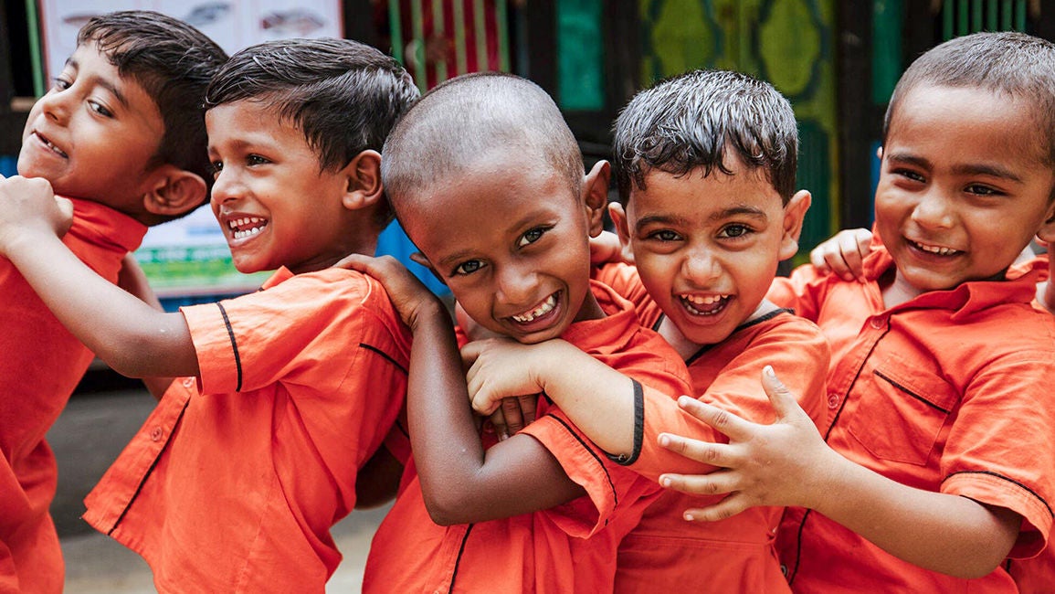 Three young boys wearing orange uniform shirts are hugging each other and smiling.