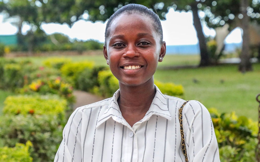 A young woman wearing a striped white shirt and standing in a manicured garden smiles.